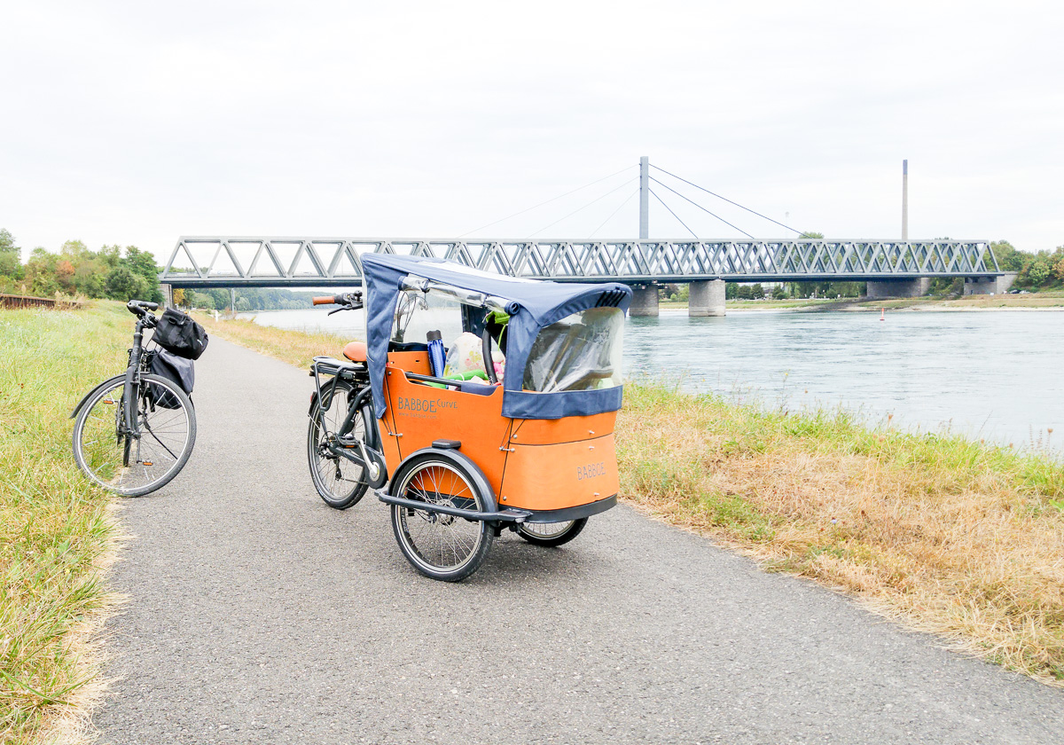 Brücke über dem Rhein Babboe Lastenrad Curve-E Elektro Lastenfahrrad für Fahrradurlaub und Fahrradtour mit Baby und Kind vorne drin 